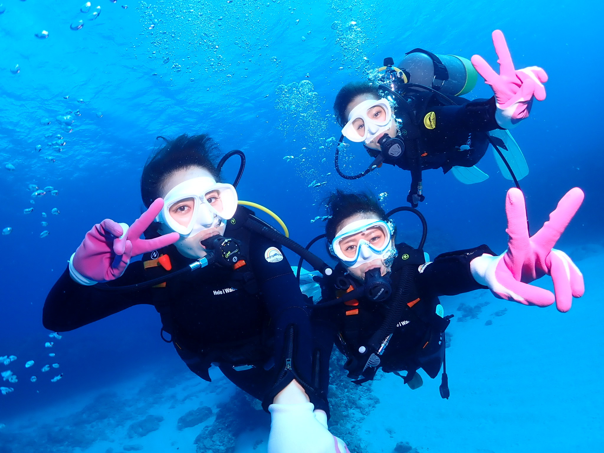 写真：ダイビング中の3人の女性。海中の青を背景に白いマスクとピンクの手袋でVサインをしている。