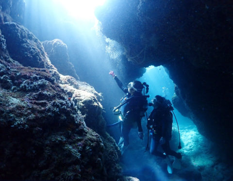 写真：海底の岩の隙間に差し込む青白い光と2人のダイバー